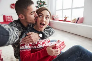 Man and woman dressed in Christmas clothing hugging and holding a gift